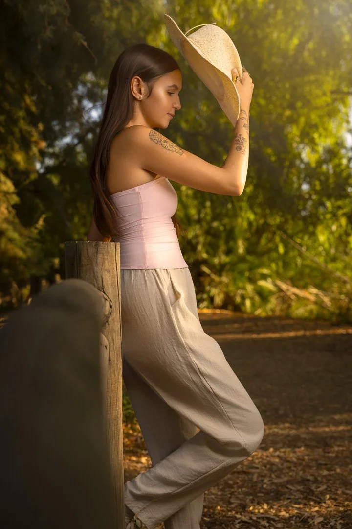 Una mujer joven con cabello largo y oscuro, con tatuajes en el brazo, sosteniendo un sombrero de ala ancha y mirando hacia abajo, de pie al aire libre en un entorno natural con árboles y un camino.