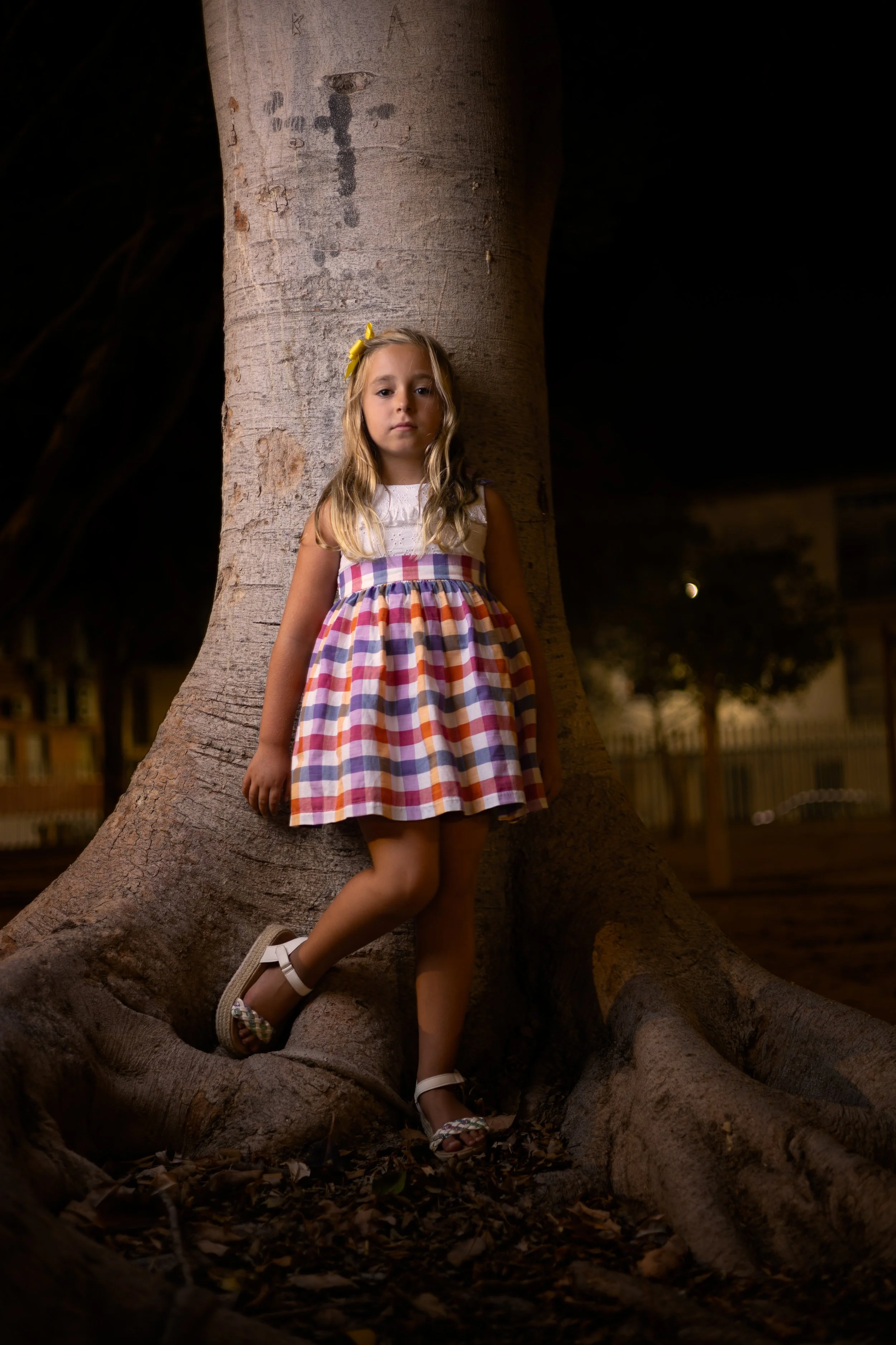 Niña con vestido a cuadros, cabello rubio y suelto, con lazos amarillos, apoyada contra un árbol en la noche.