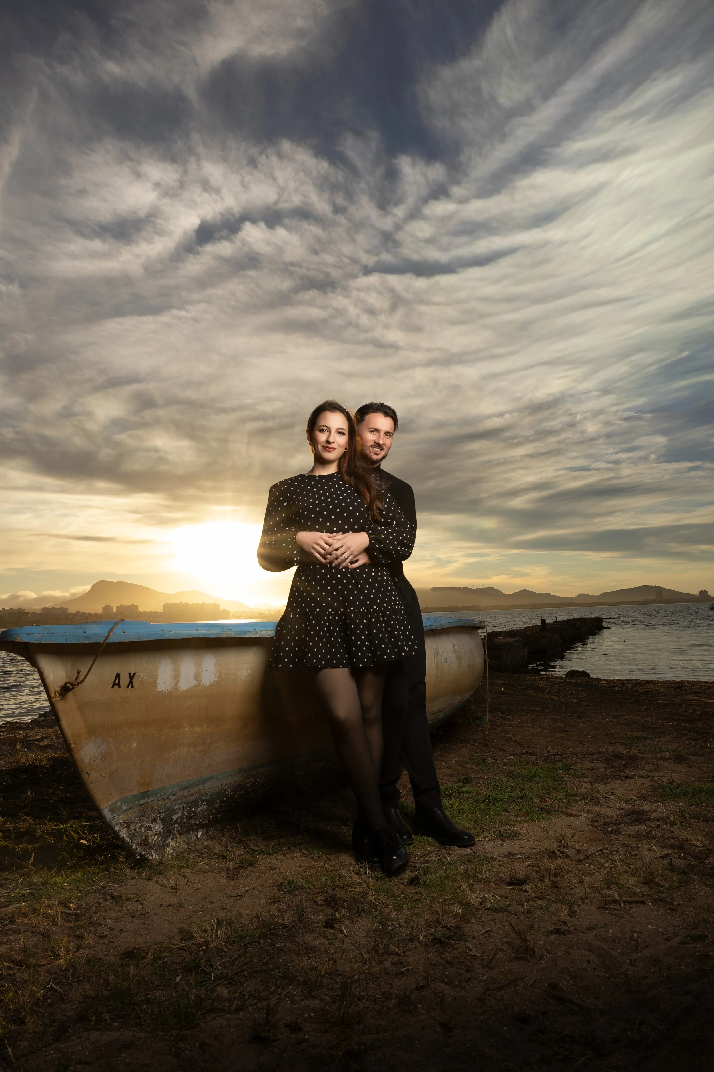 Una pareja de pie junto a un barco en la orilla, con el sol poniendo atrás y un cielo con nubes.