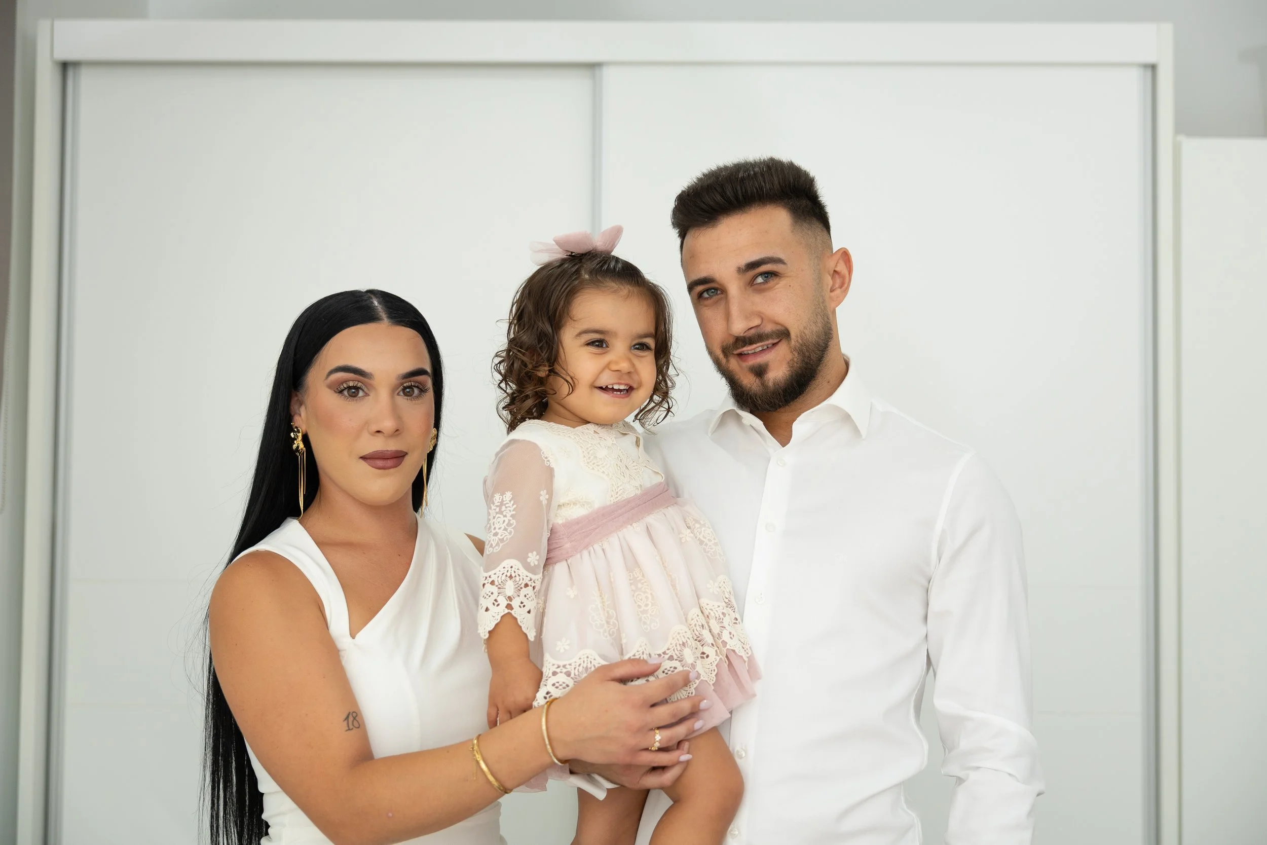 Una familia feliz con una mujer, un hombre y una niña pequeña posando para la cámara, todos vestidos con ropa blanca, en un ambiente interior con fondo blanco.