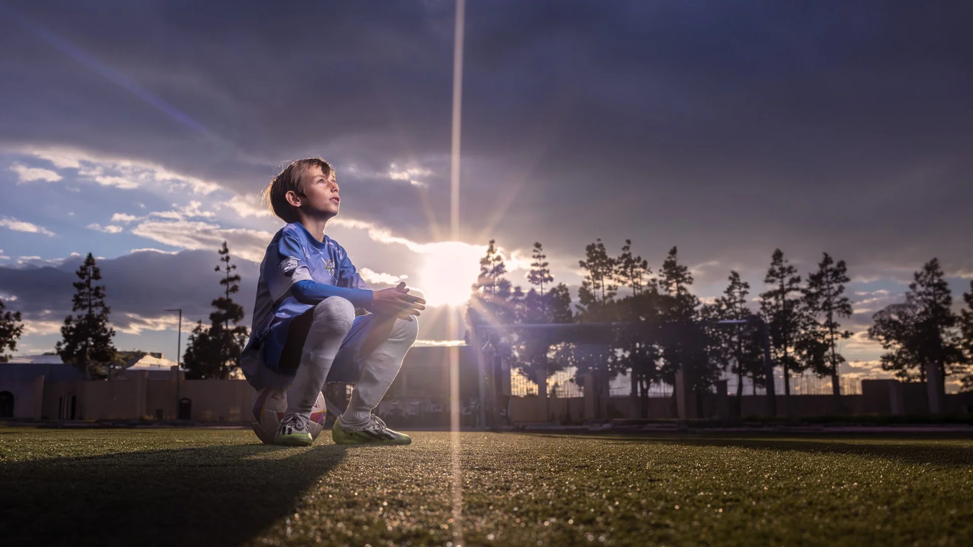 Niño con uniforme de fútbol sentado en el campo con el sol poniente y árboles en el fondo.