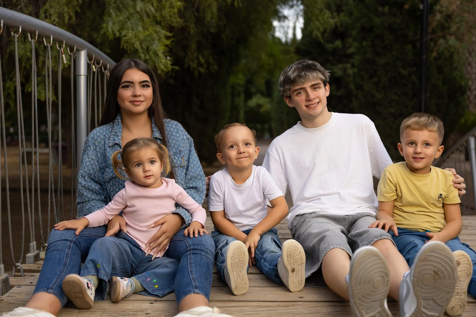 Una familia de cinco personas sentadas en un puente de madera en un parque, rodeados de árboles verdes.