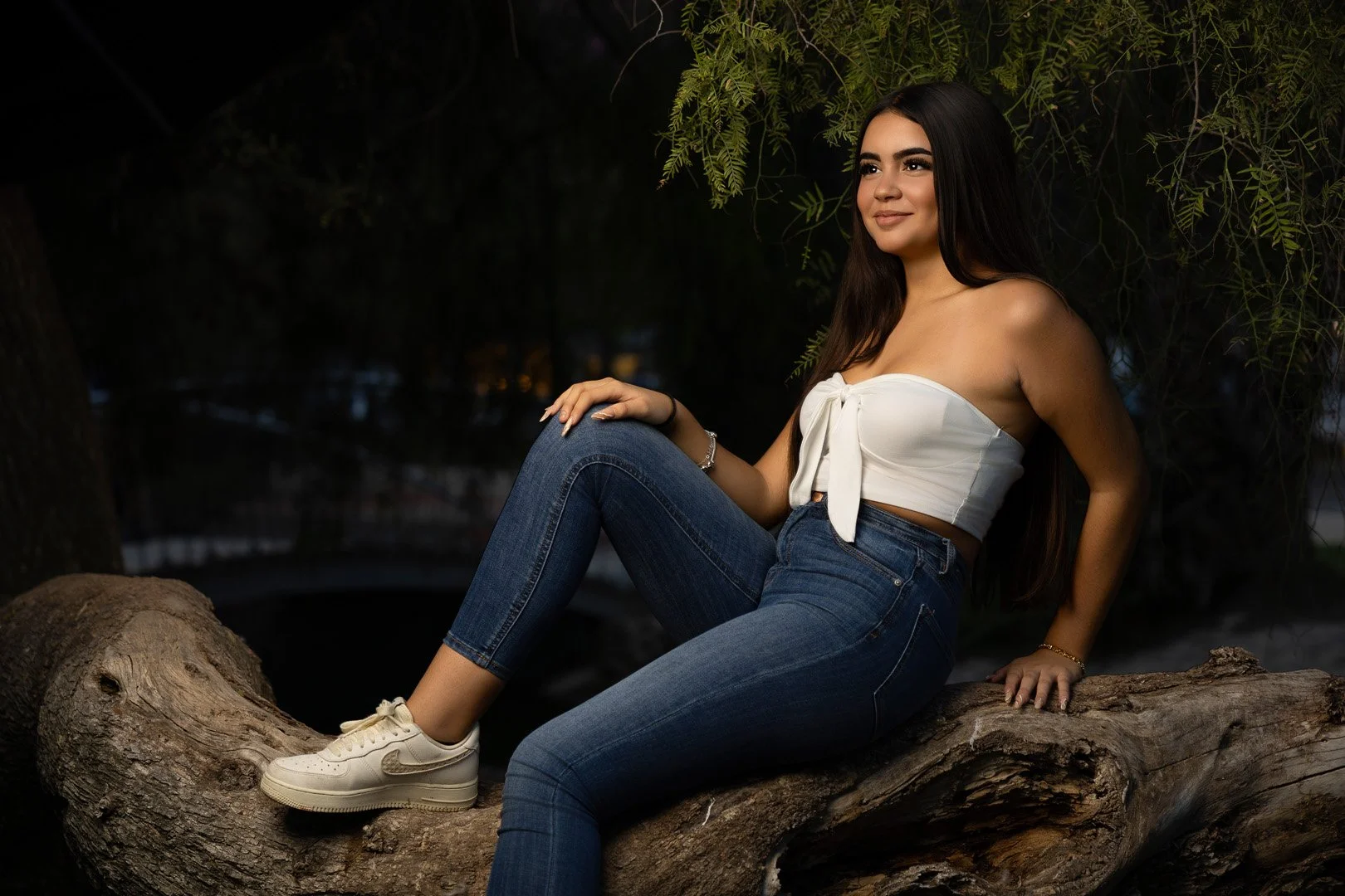Joven mujer sentada en una rama de árbol en un entorno nocturno, con cabello largo y oscuro, usando blusa blanca con lazo, jeans azules y zapatillas blancas.