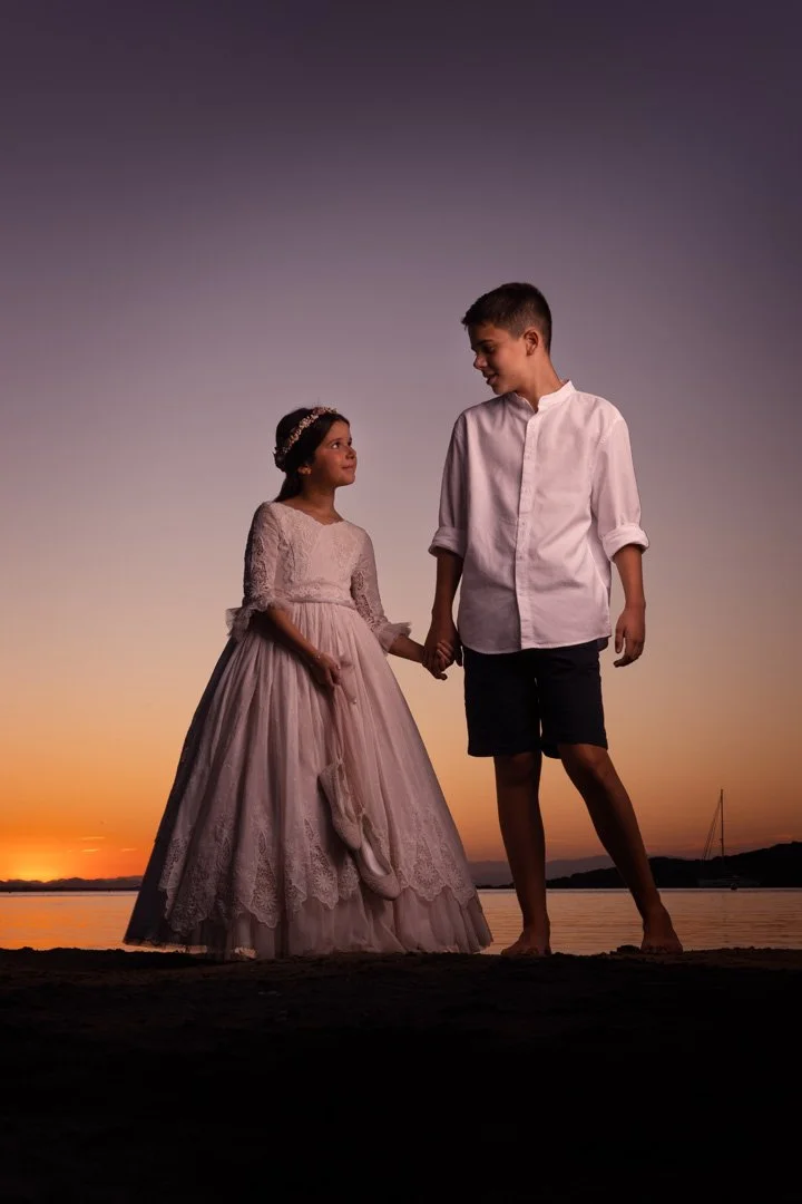 Un niño y una niña adolescentes en la playa al atardecer, la niña viste un vestido de fiesta y el niño lleva una camisa y pantalones cortos, ambos se toman de las manos y se miran con cariño.