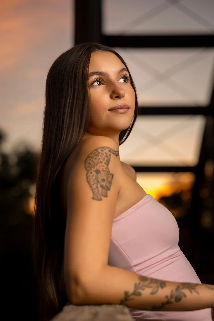 Joven mujer con tatuajes en el brazo y el hombro, cabello largo y oscuro, mirando al cielo durante el atardecer, usando un top rosa.