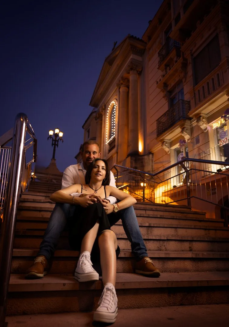 Una pareja sentada en las escaleras de una calle iluminada por luces de noche, con edificios y farolas en el fondo.