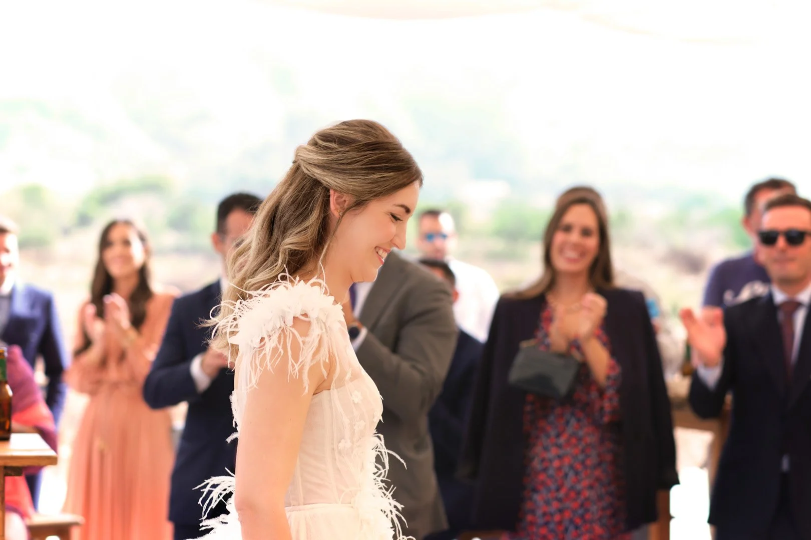 Una mujer sonriente en vestido de novia en una ceremonia con invitados en el fondo.