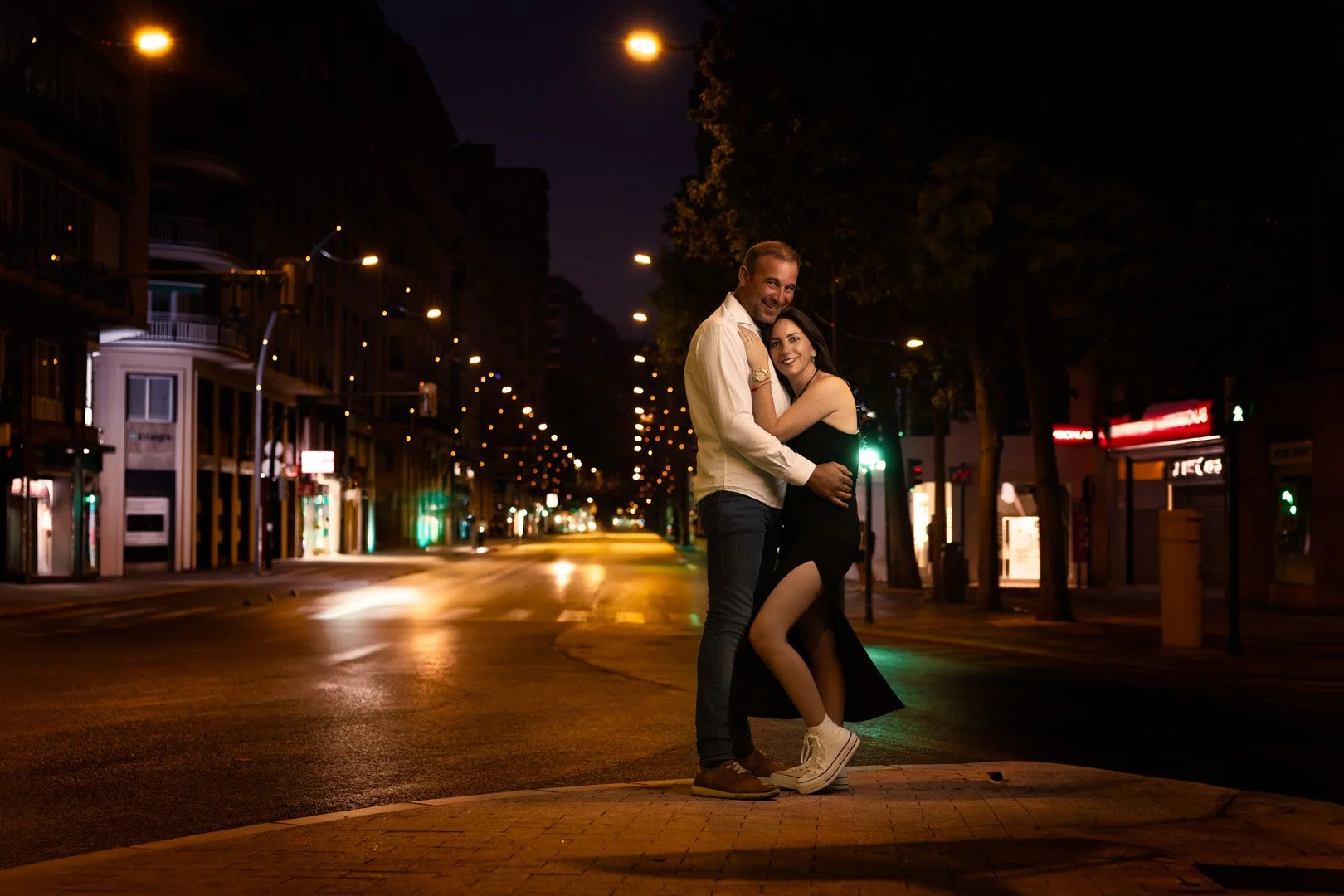 una pareja abrazada en una calle vacía y iluminada por farolas durante la noche.