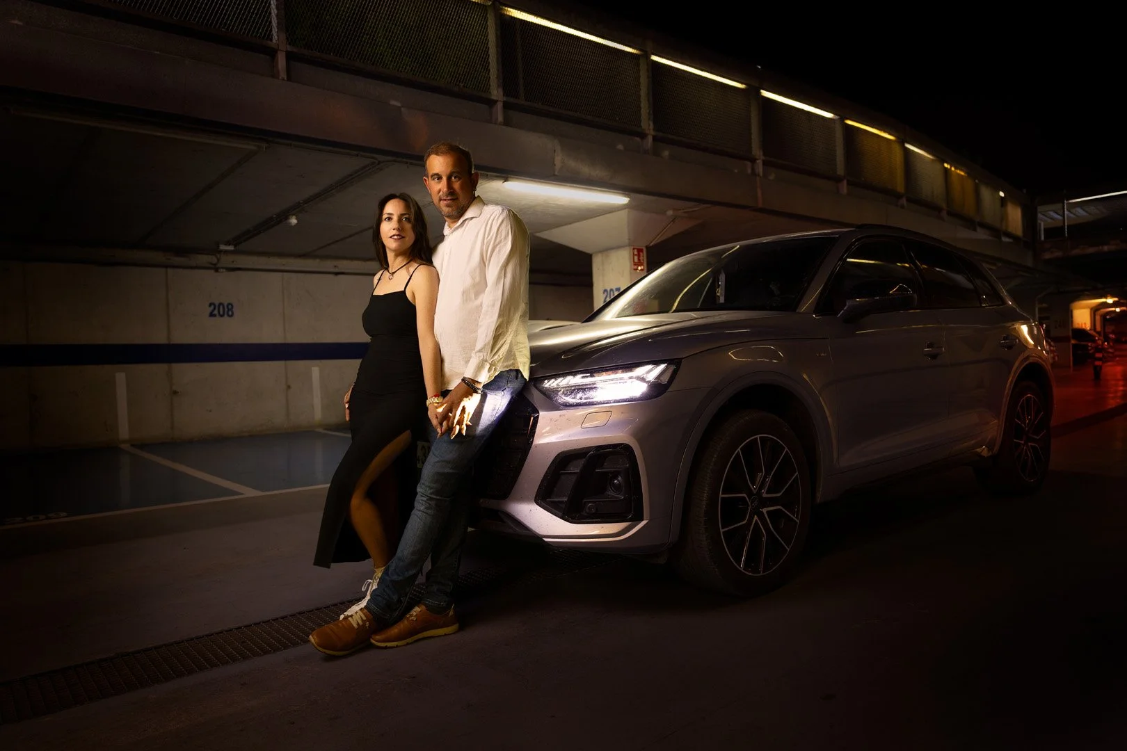 Una pareja posa junto a un coche gris oscuro en un estacionamiento subterráneo de noche.