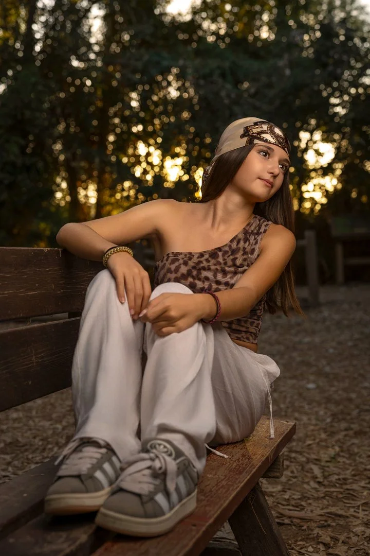 Joven sentada en un banco en un parque, vista lateral, con cabello largo, vestido de estilo casual con camiseta de animal print, pantalones blancos y zapatillas deportivas, durante el atardecer con fondo de árboles y luz dorada.