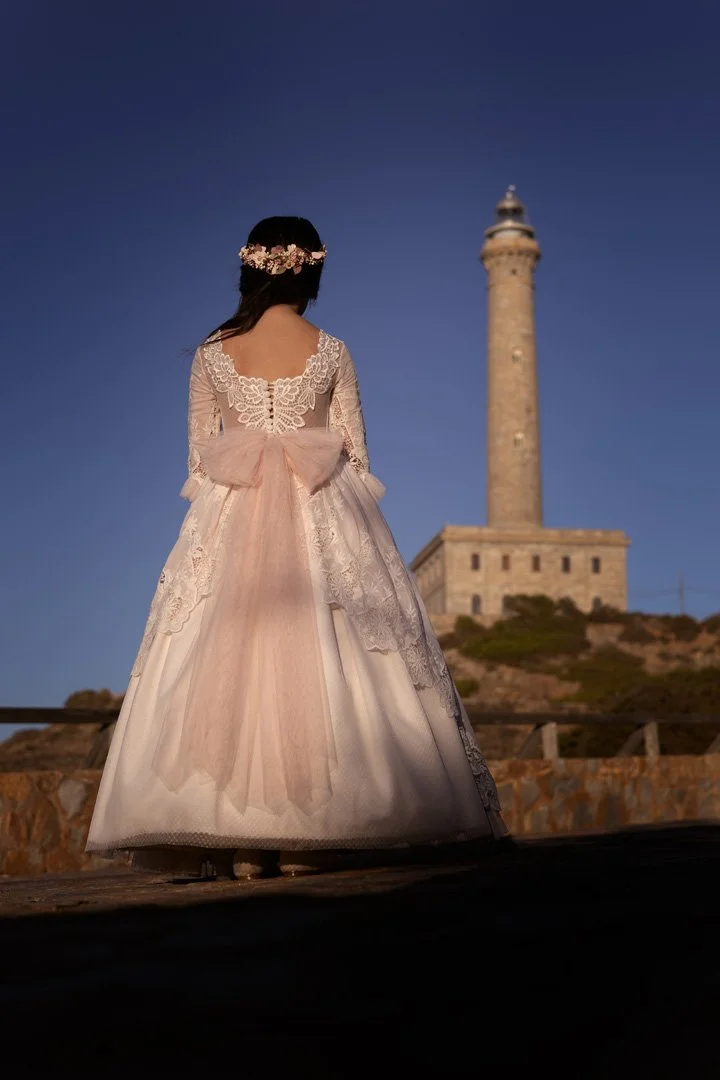 Mujer vestida con un vestido largo y liso con encaje y un lazo de tul en la espalda, usando una corona de flores, de espaldas, mira hacia una torre en una colina bajo un cielo azul.
