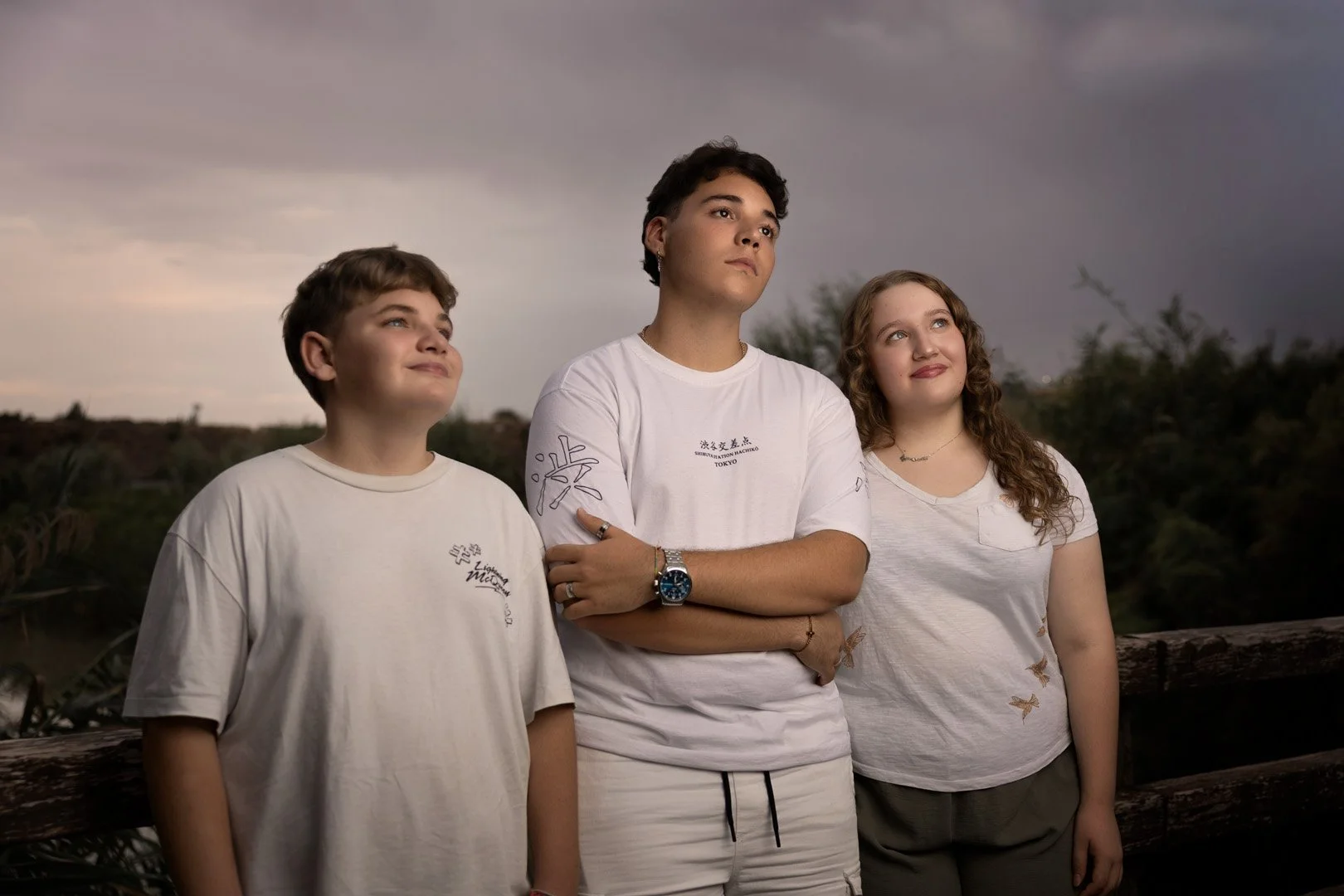 Tres jóvenes frente a un fondo natural con nubes, mirando pensativos en la tarde o noche.