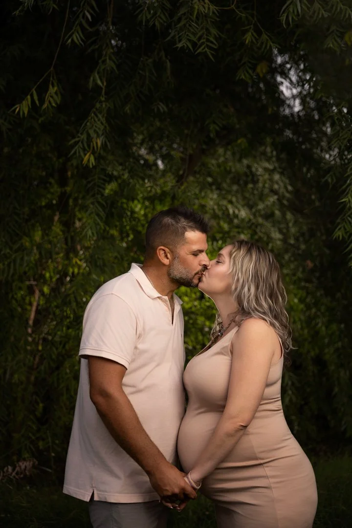 Pareja embarazada besándose en un entorno natural con árboles y follaje.