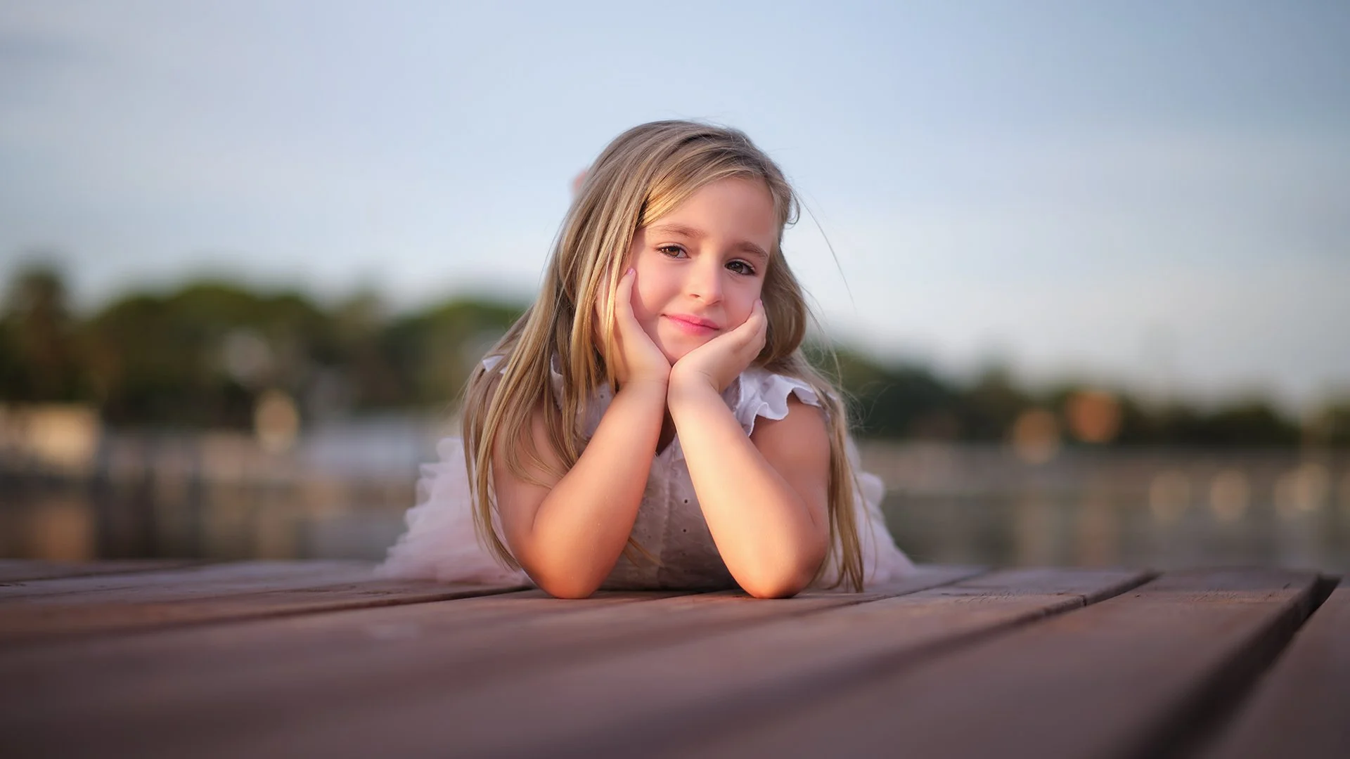 Niña rubia acostada en un muelle de madera, con expresión pensativa, con fondo de paisaje natural y agua.