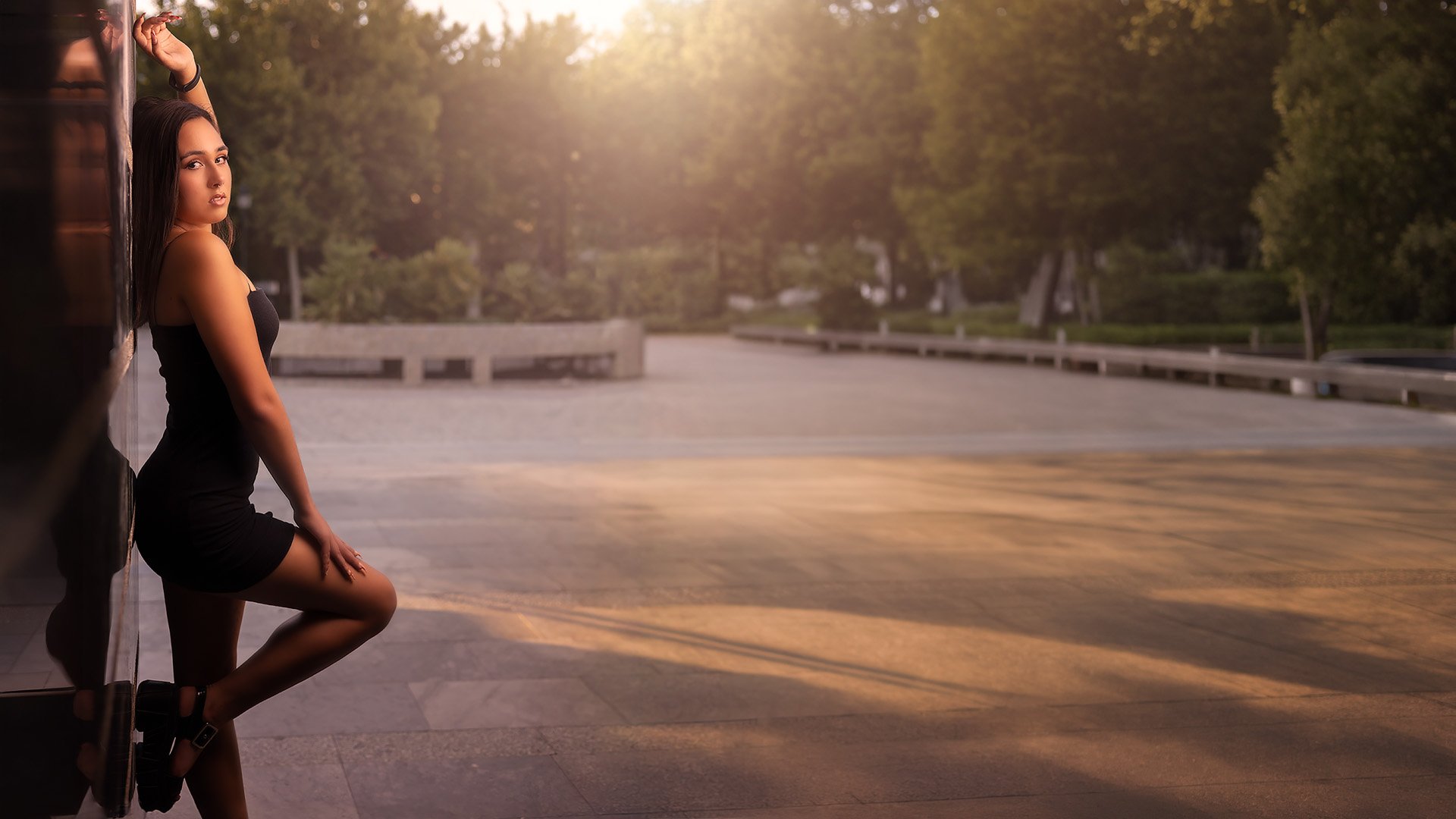 Mujer con vestido negro posando en la esquina de un edificio, con un fondo de calles y árboles al atardecer.