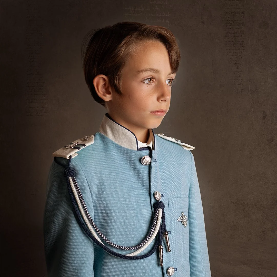 Joven en uniforme azul con insignia militar y cordón decorativo, de cabello castaño claro, mirando hacia la derecha con expresión seria, en fondo neutro.