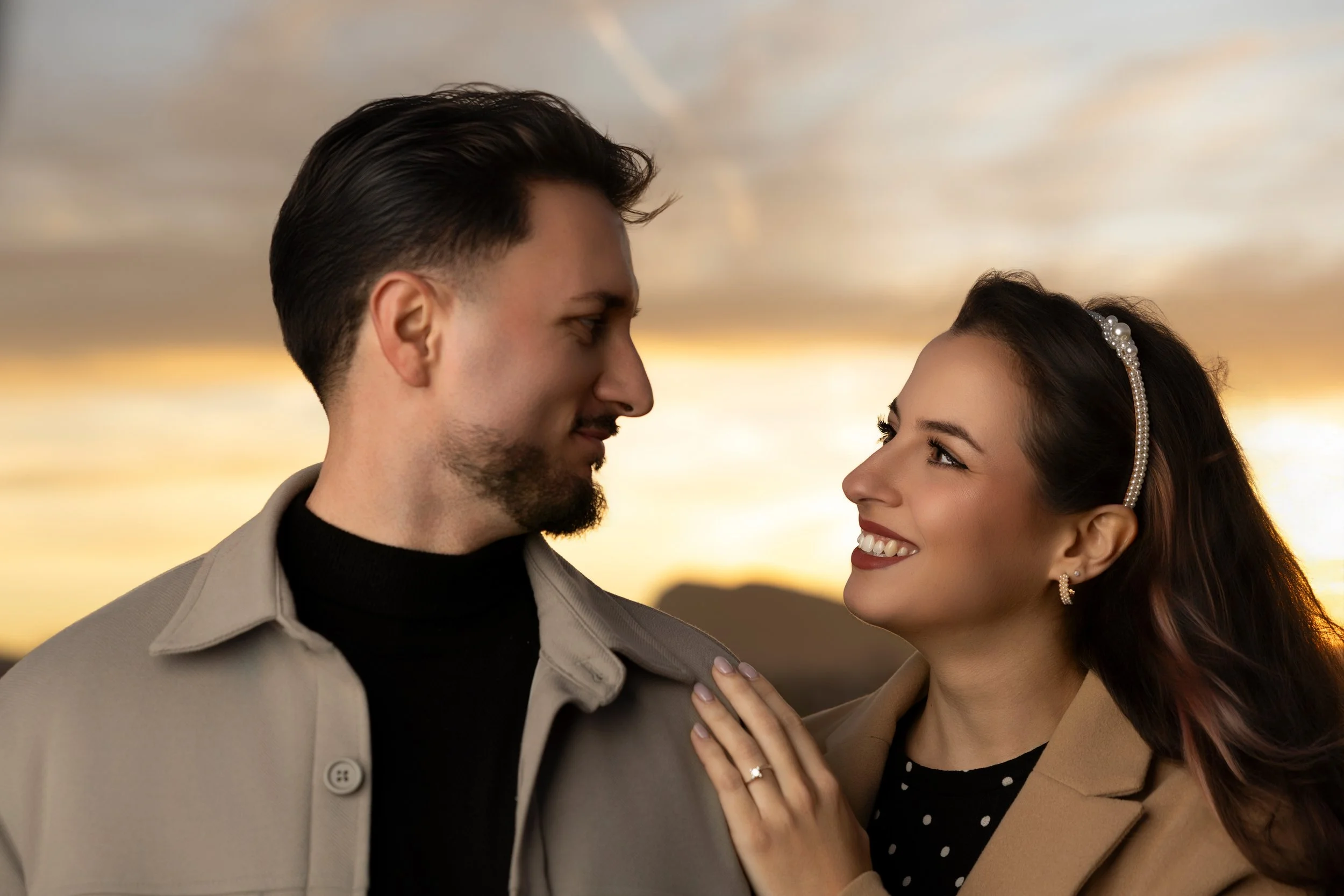 Una pareja mirándose con amor al atardecer, la mujer sonriente con accesorios de perlas en el cabello y el hombre con barba y cabello oscuro.