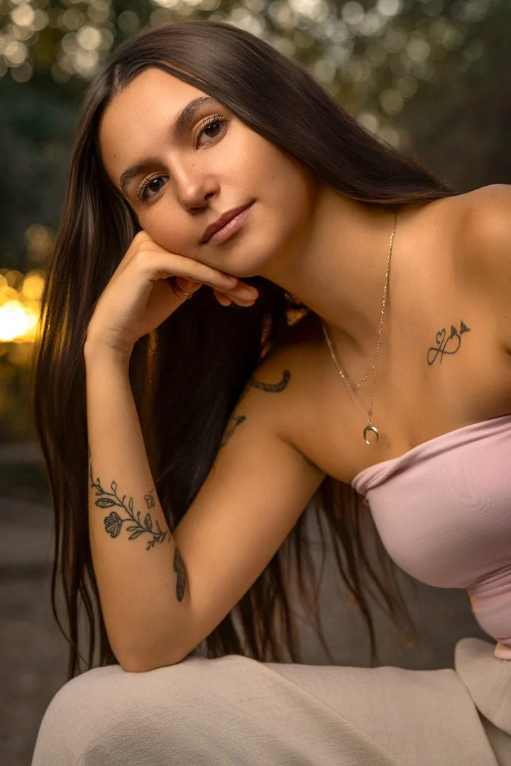 Una joven con cabello largo y oscuro, mirando a la cámara con una expresión tranquila, apoyada en su mano, con tatuajes en el brazo y el pecho, usando un collar, en un entorno natural al atardecer.