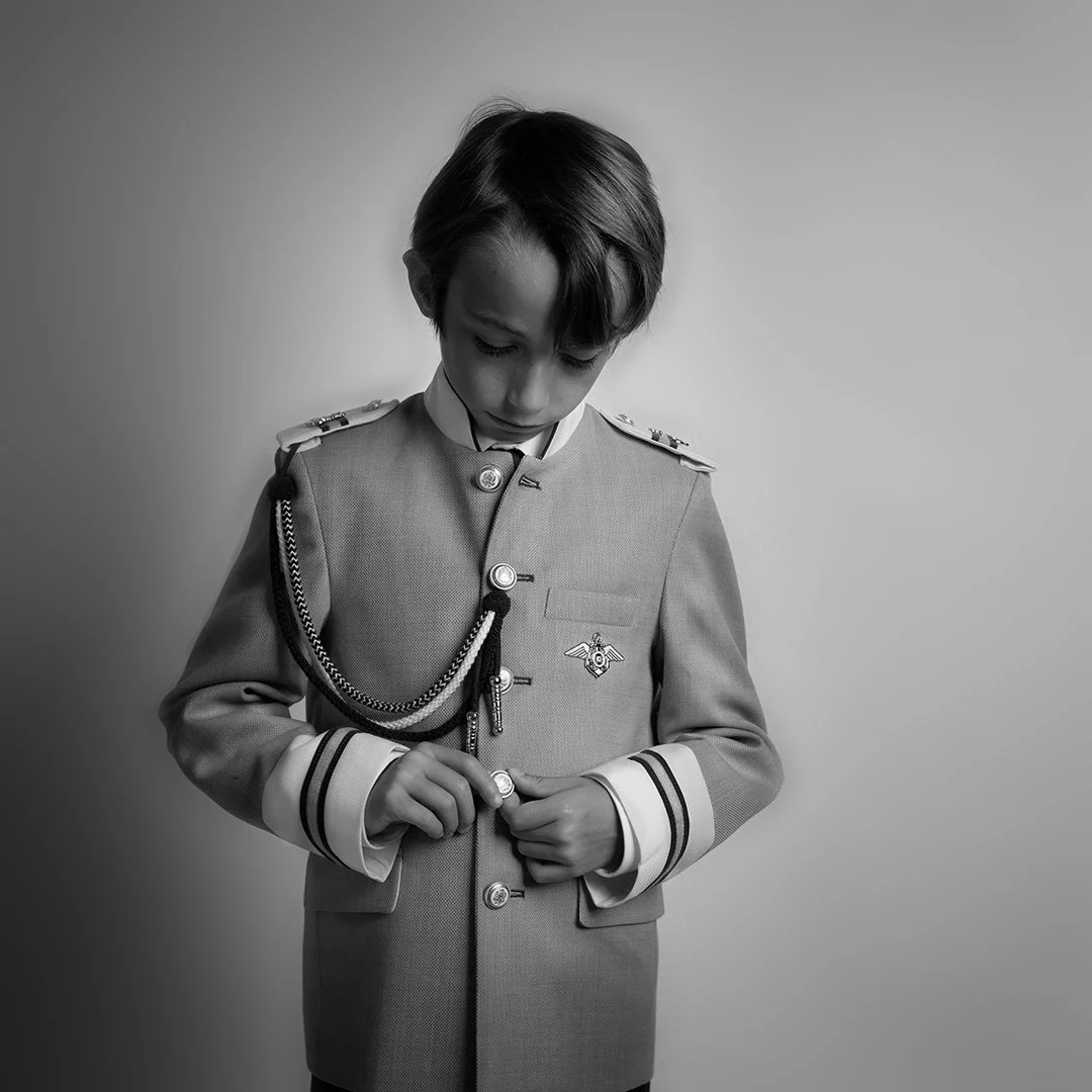 Retrato en blanco y negro de un joven niño con uniforme militar, ajustando un botón en la parte frontal de su chaqueta.