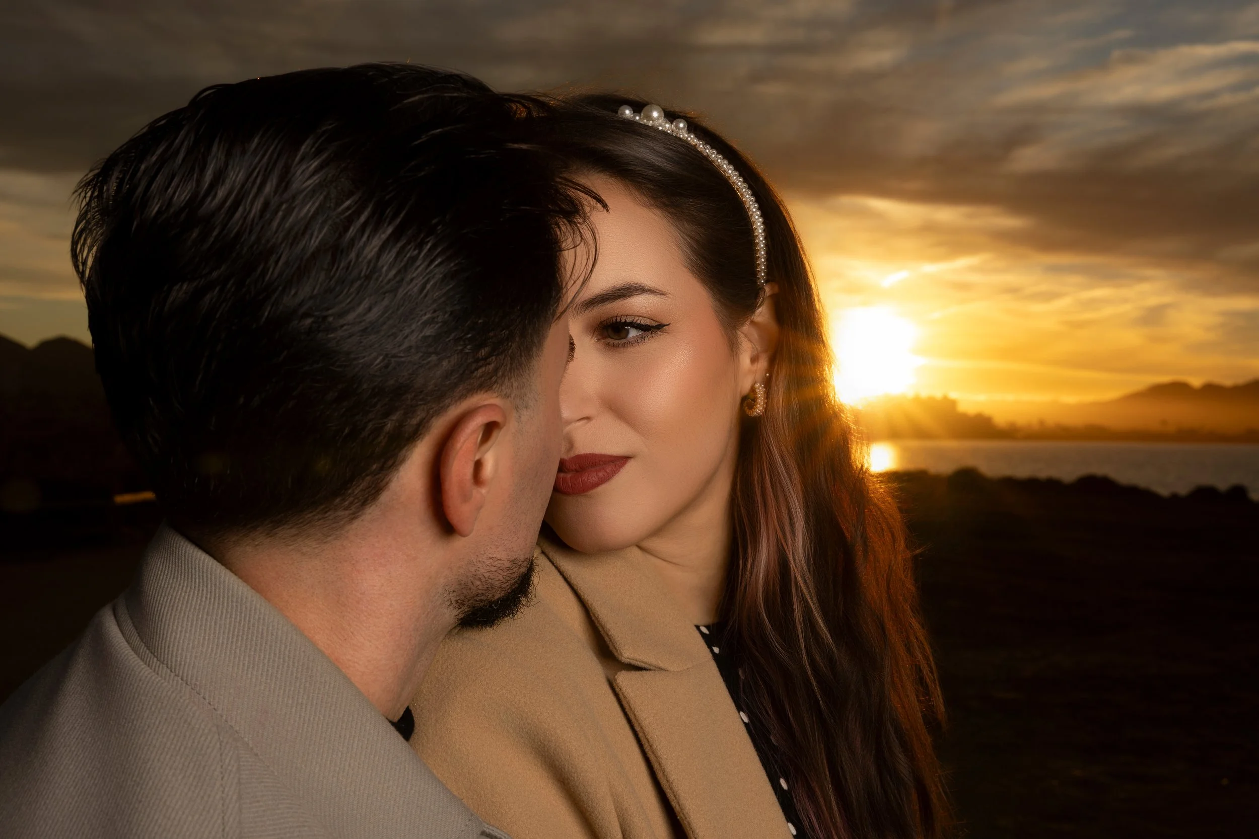 Una pareja en un abrazo cercano al atardecer, con el sol iluminando el paisaje y las montañas al fondo.