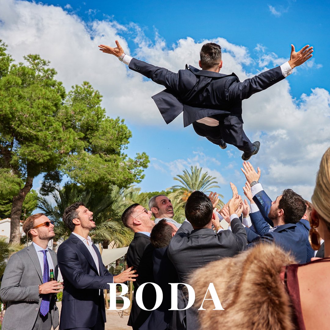 Grupo de personas vestidas con ropa formal celebrando al lanzar a un hombre al aire en una ceremonia de boda en un parque con árboles y cielo azul.