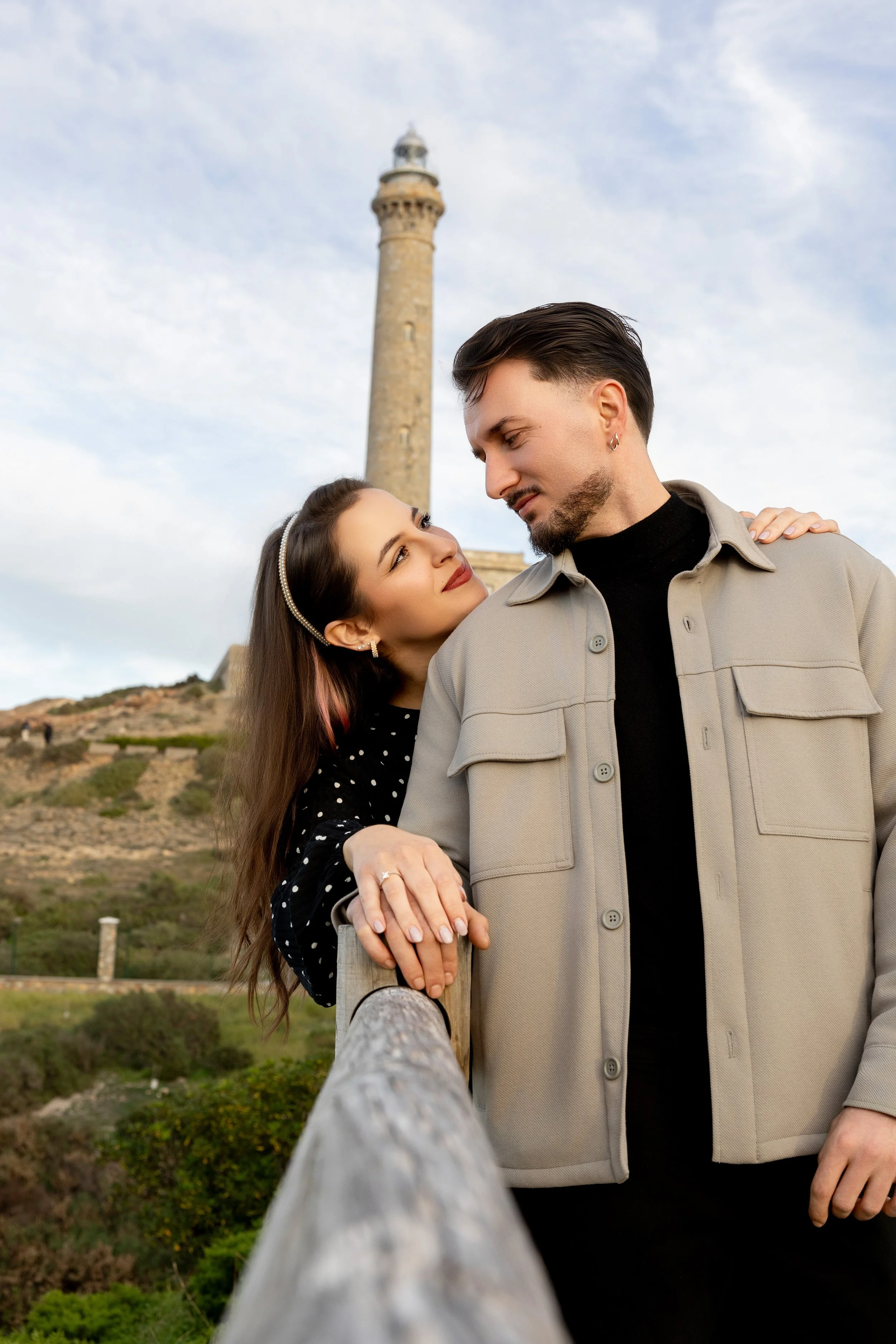 Una pareja mira amorosamente en un mirador con un faro en el fondo, en un día con cielo despejado.