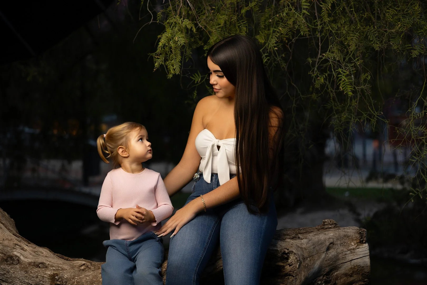 Una mujer joven y una niña pequeña están sentadas en un tronco de árbol en un entorno natural al atardecer, conversando y mirando atentamente la una a la otra.