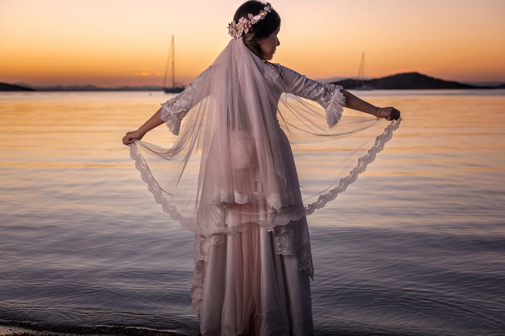 Una mujer vestida con ropa de época en una orilla de río durante el atardecer, sosteniendo un velo con encaje.