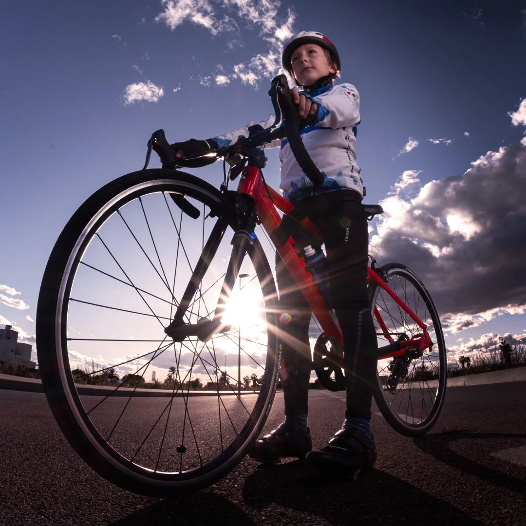 Niño con casco y ropa deportiva montando una bicicleta roja en una carretera con el sol detrás y un cielo nublado.