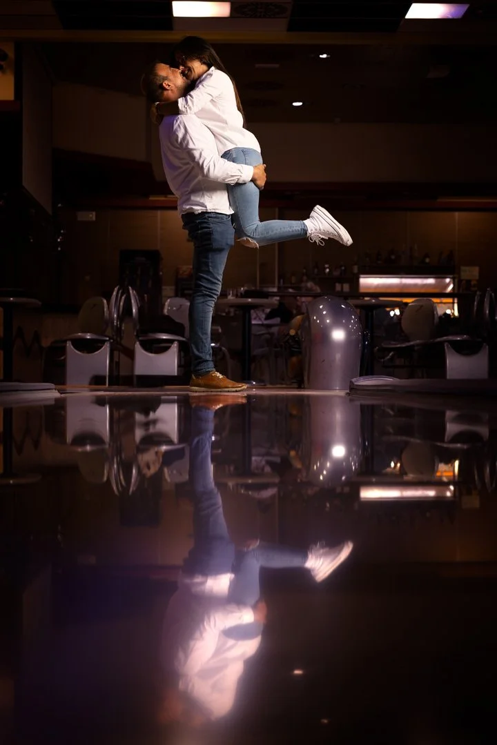Una pareja en un bando, la mujer levitando en el aire y siendo abrazada por el hombre en un ambiente de salón con sillas y mesas, reflejo en el suelo.