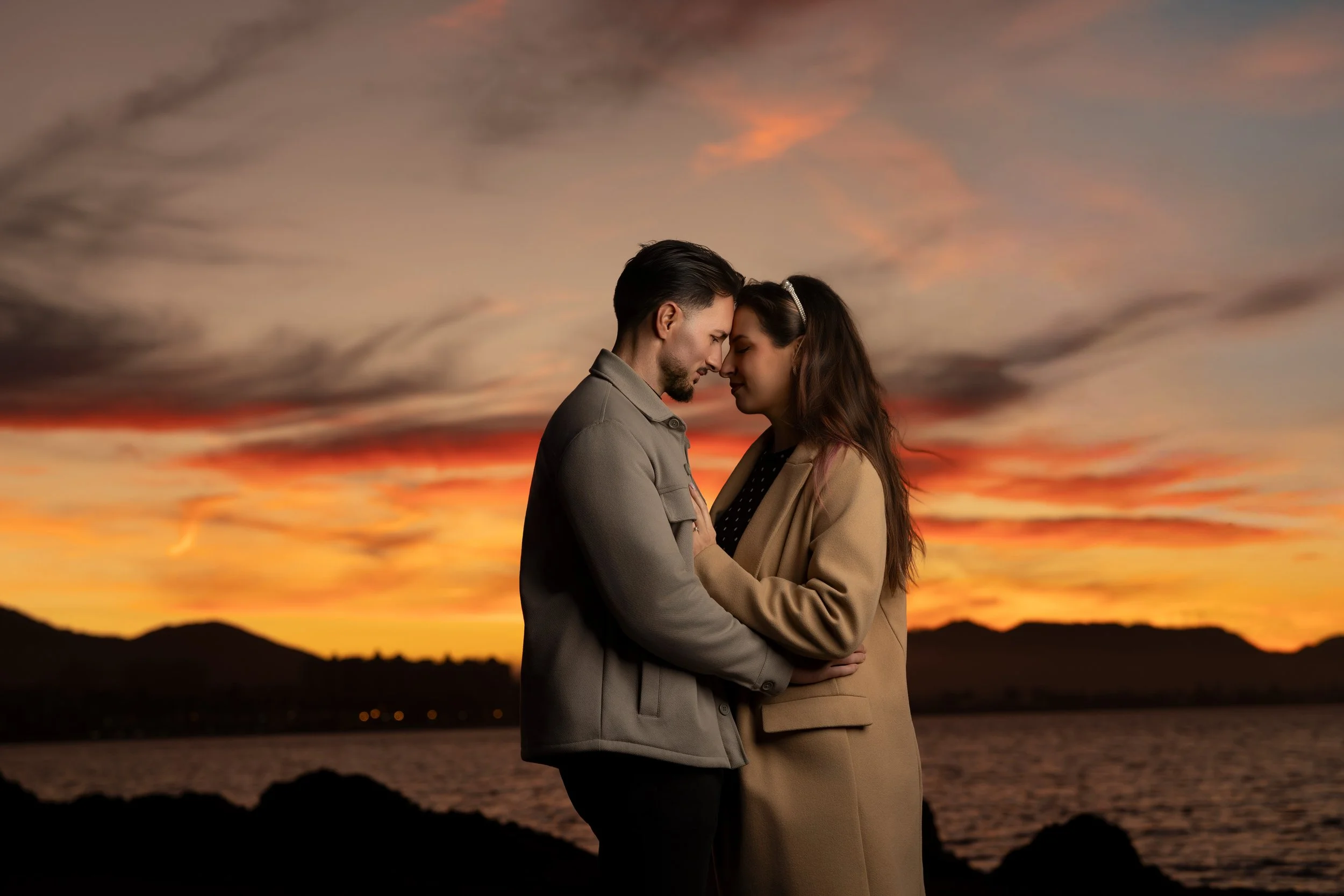 Una pareja se abraza y toca sus frentes en un atardecer con cielo colorido sobre un lago y montañas.