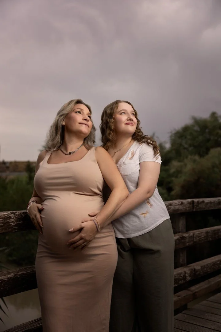 Dos mujeres, una embarazada, abrazadas y mirándose hacia arriba en un puente, con cielo nublado de fondo.