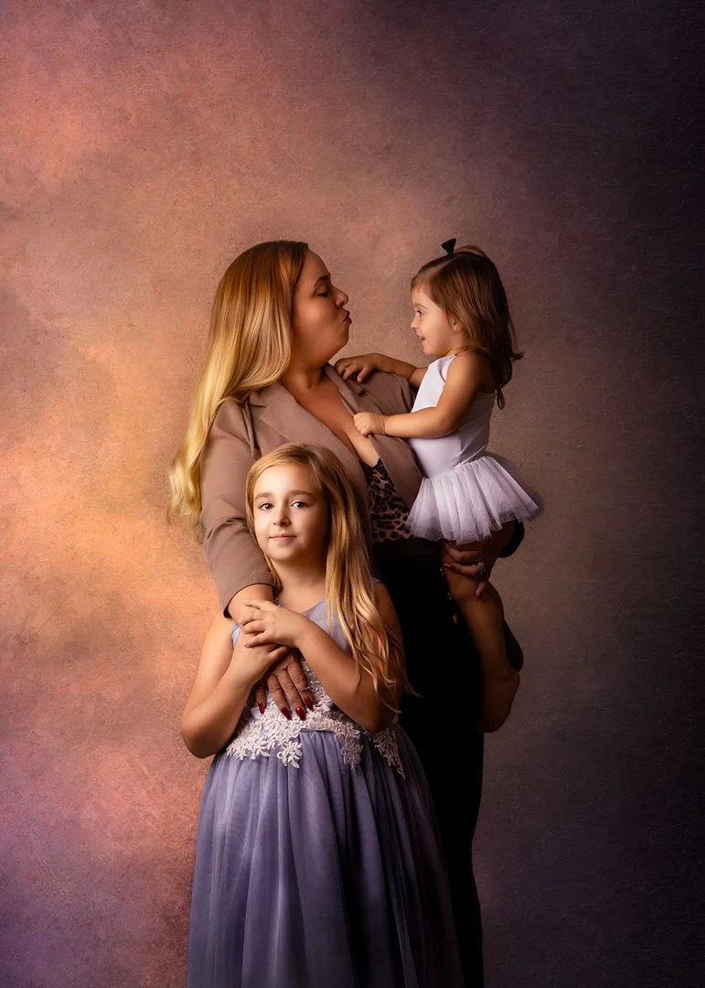 Mujer con dos niñas pequeña y adolescente en un retrato familiar, la mujer sostiene a una niña más joven mientras otra niña la abraza por la cintura.