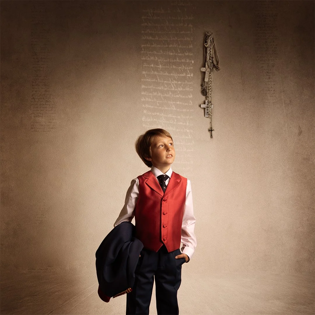Un niño con saco rojo, camisa blanca, y corbata, sosteniendo una chaqueta oscura en la mano, mirando hacia arriba, en una pared con crucifijos y rosarios colgados.