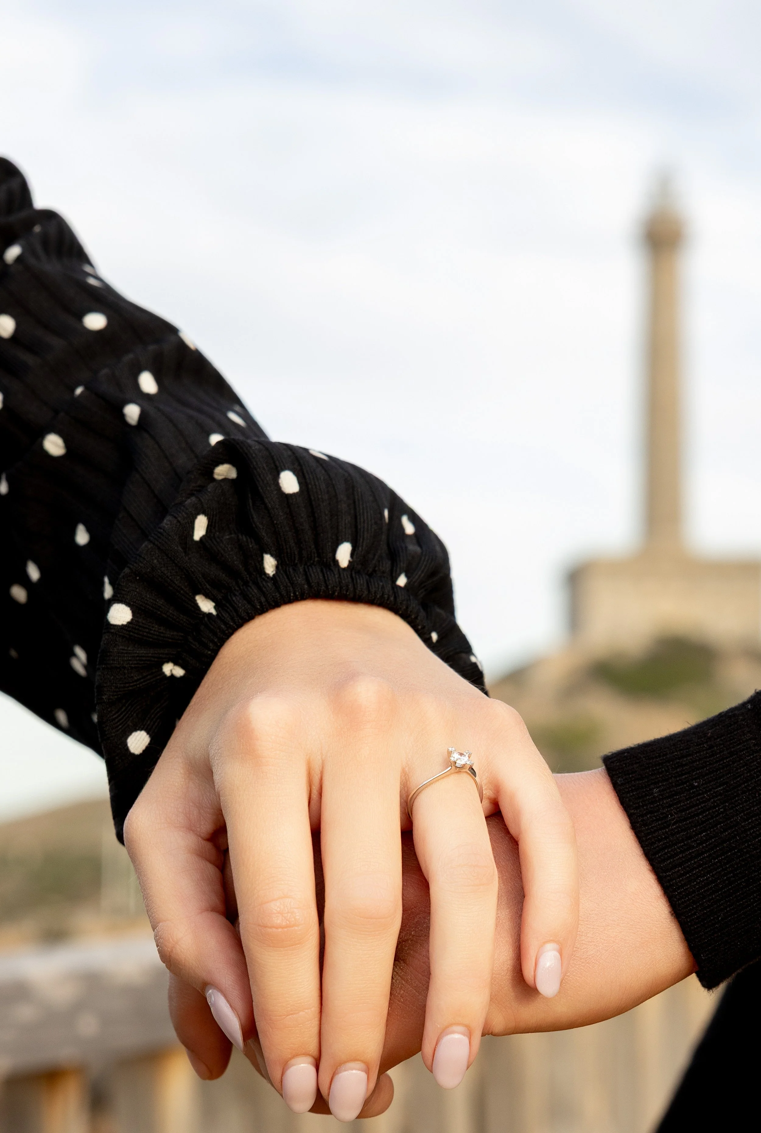 Primer plano de dos manos entrelazadas con anillo de compromiso con diamante en el dedo de la mujer, con fondo de un monumento y cielo despejado.