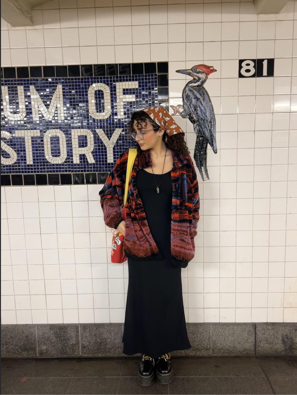 isa benkhallouk stands beside the "museum of natural history" subway stop. she has curly hair and glasses. in her hair is an orange scarf with cats illustarted in it. her hand is in her colorful printed coat pocket and she's looking off to the side.