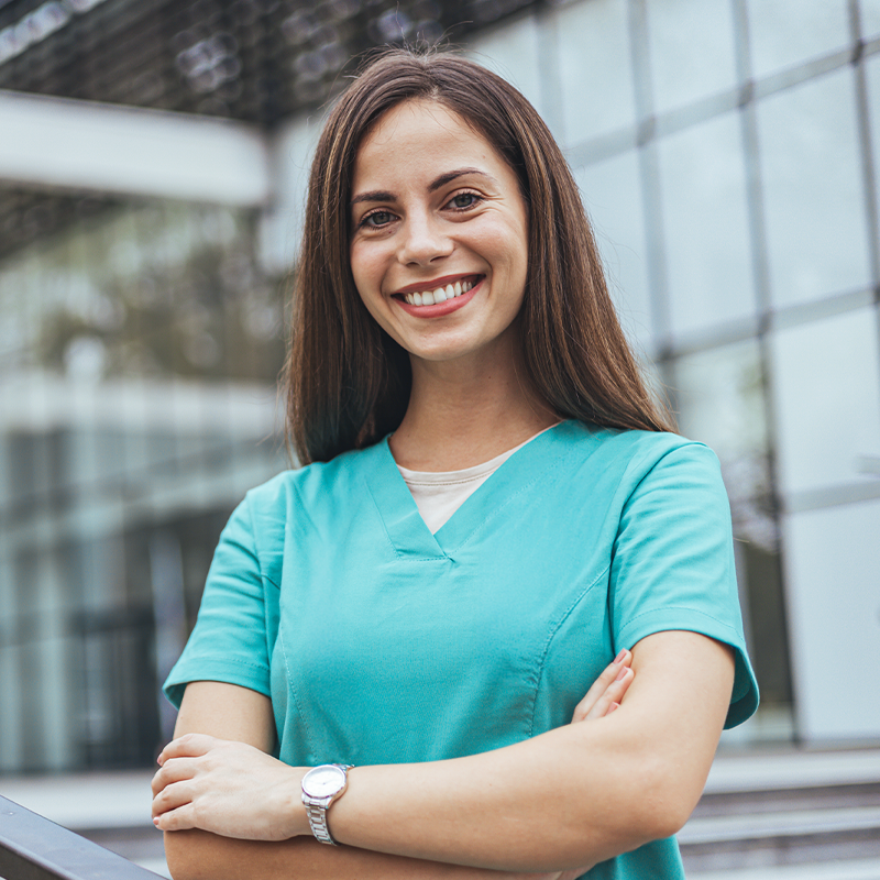 A smiling woman in teal medical scrubs standing outside a modern glass building with her arms crossed.