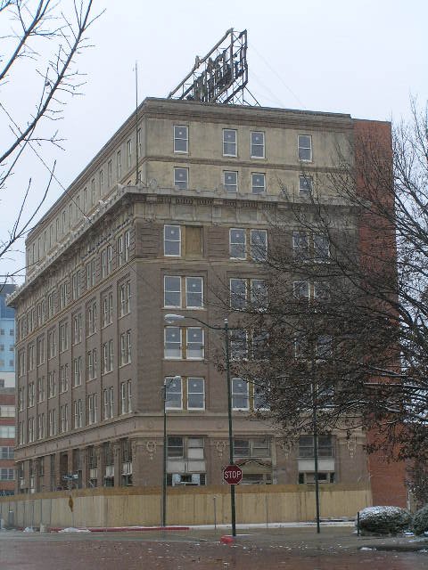 A multi-story building with a billboard structure on the roof, surrounded by leafless trees and a construction fence at the base, in an urban setting.