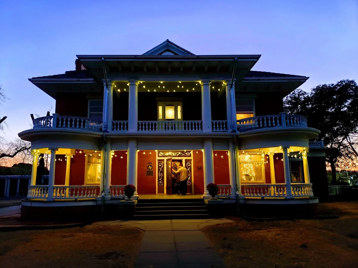 A large, two-story Victorian-style house decorated with string lights, illuminated at dusk, with a person standing at the front door.