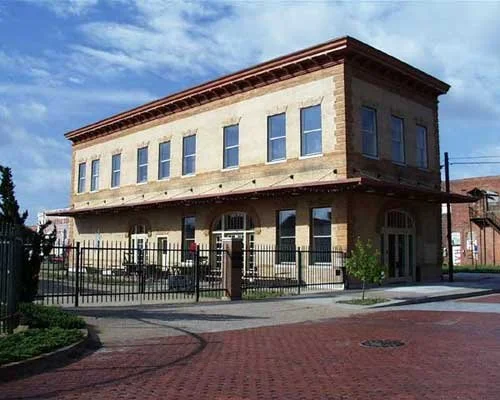 Two-story brick building with a fenced patio, set on a brick-paved street, under a partly cloudy sky.