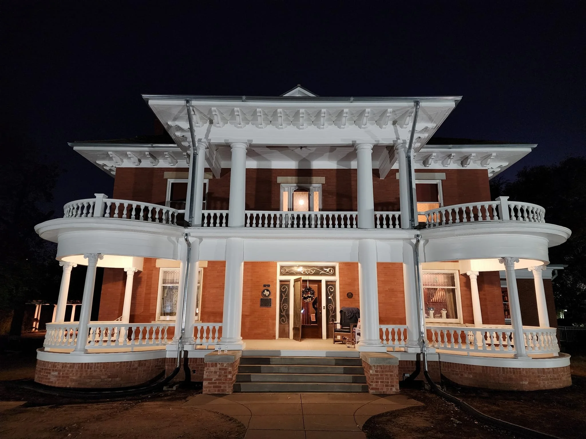Night view of a large, white, three-story Victorian-style house with detailed trim, columns, and a wraparound porch accented by white balustrades.