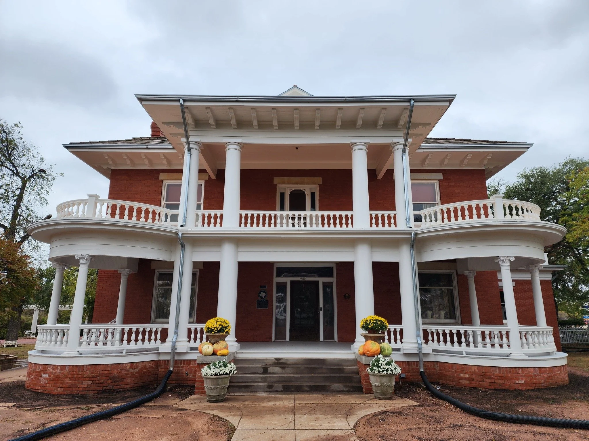 Front view of a large two-story brick house with white columns, balconies, and railings, decorated with pumpkins and flowers at the entrance, under an overcast sky.