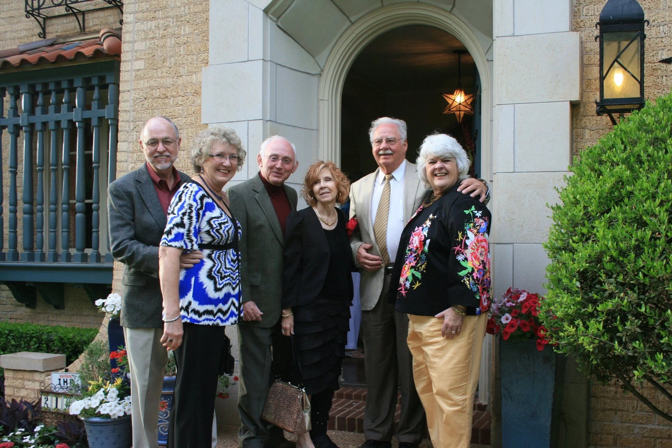 Group of seven older adults standing together in front of a house entrance, smiling for a photo, with decorative plants and flowers nearby.