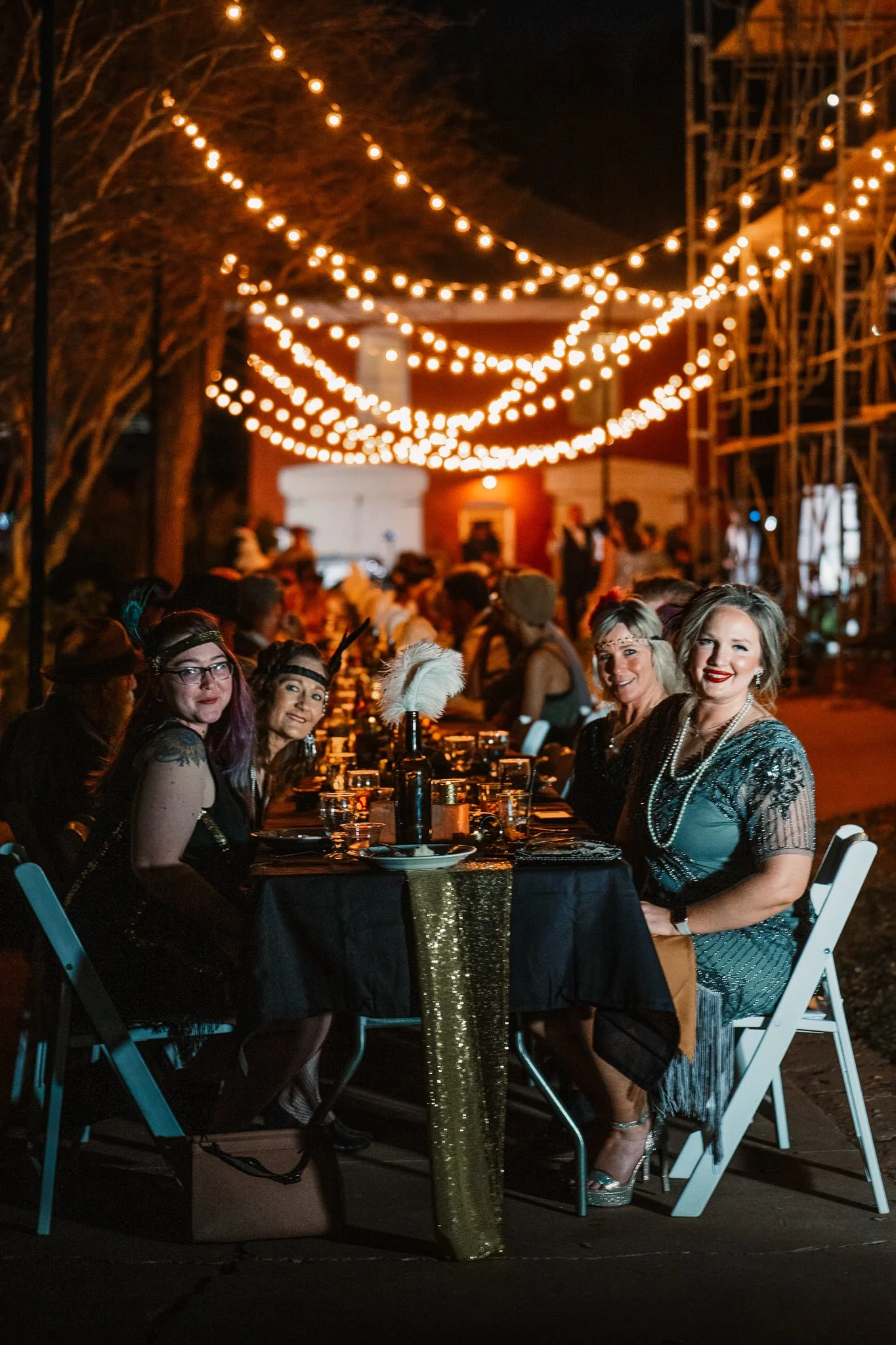 People dressed in vintage costumes gather at a long dinner table outdoors during night with string lights overhead, celebrating at a themed event or costume party.