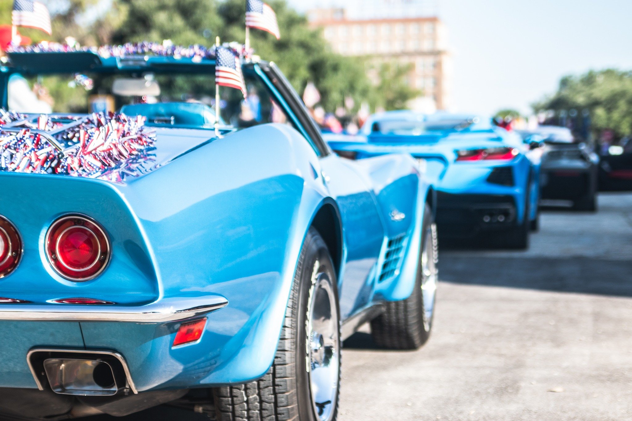 Row of blue Chevrolet Corvettes decorated for a patriotic parade with small American flags on the cars and red, white, and blue decorations in the trunk of the front car.