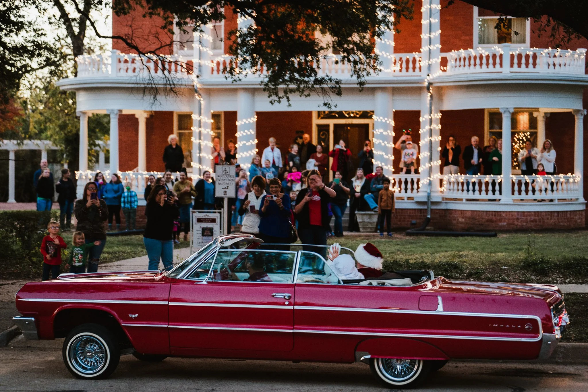 A parade featuring a person dressed as Santa Claus waving from a vintage red convertible car with people gathered on a decorated porch in the background.