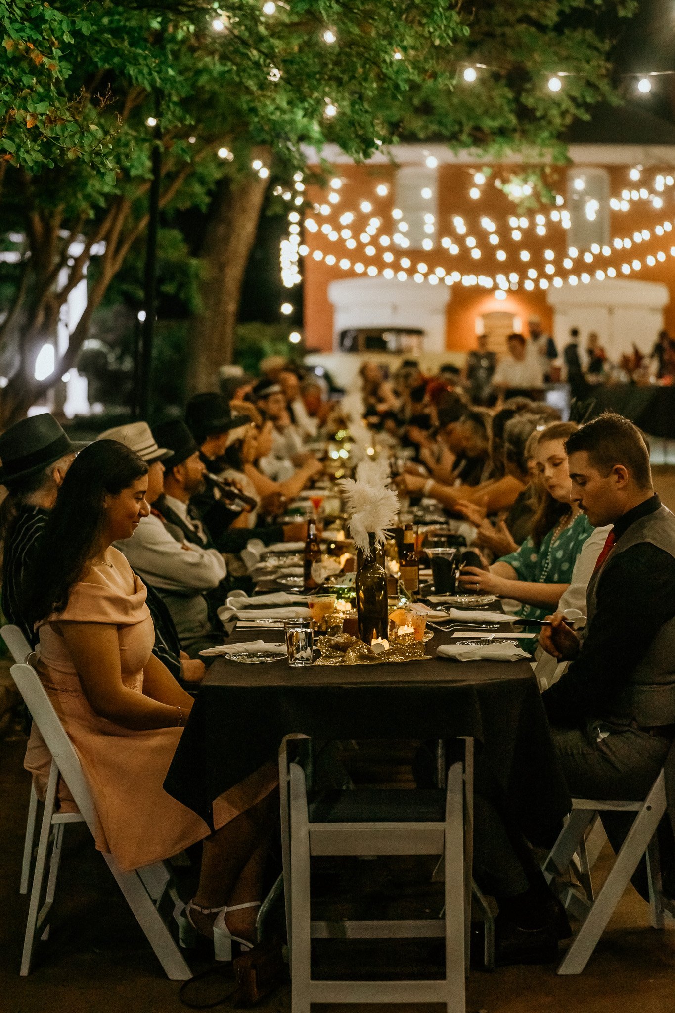 People sitting at a long outdoor dining table decorated with centerpieces and string lights during an evening event.