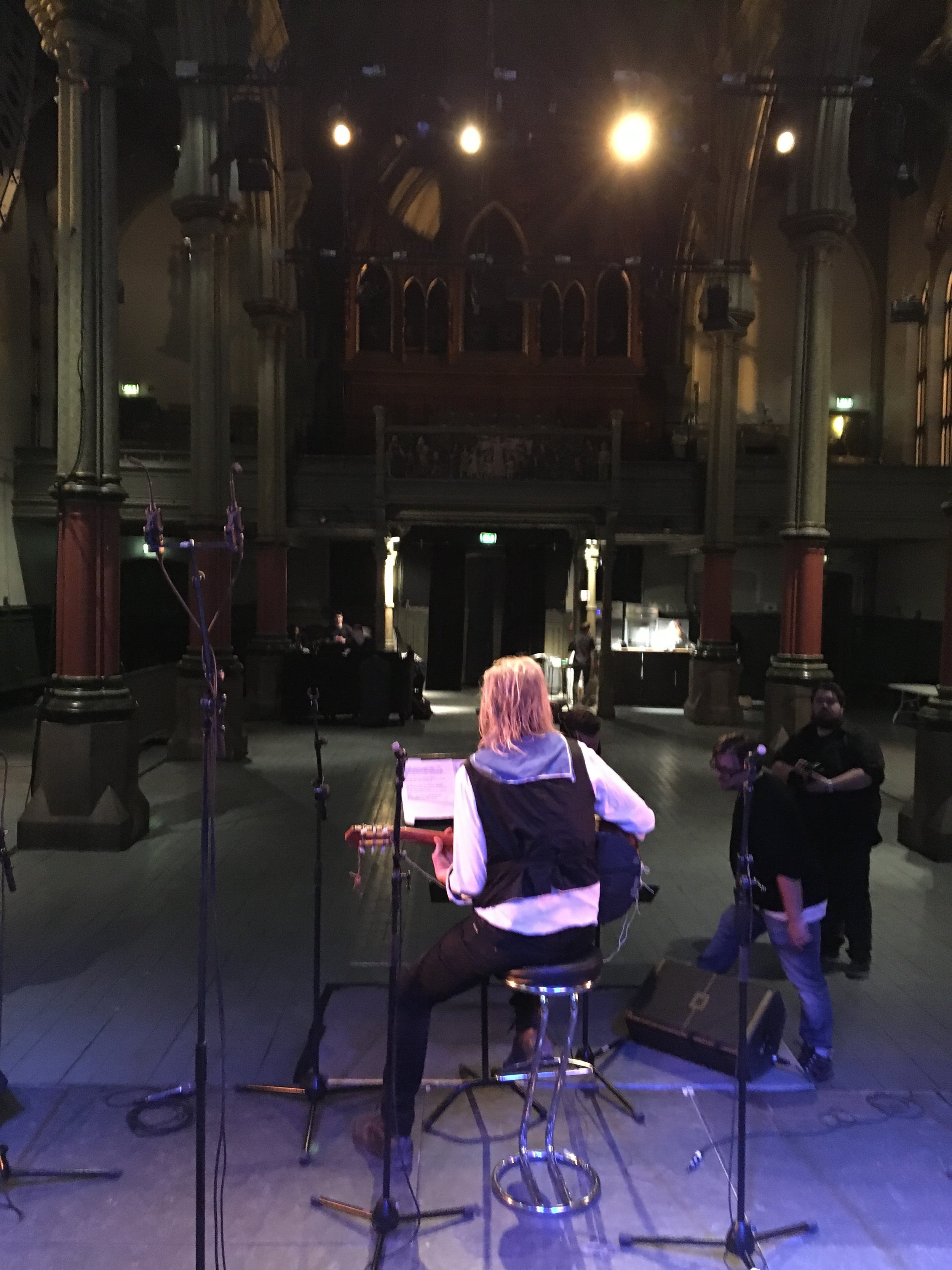 A singer with a guitar sitting on a stool on stage, with microphone stands around, in a dimly lit, historic theater or concert hall with architectural details and lighting above.