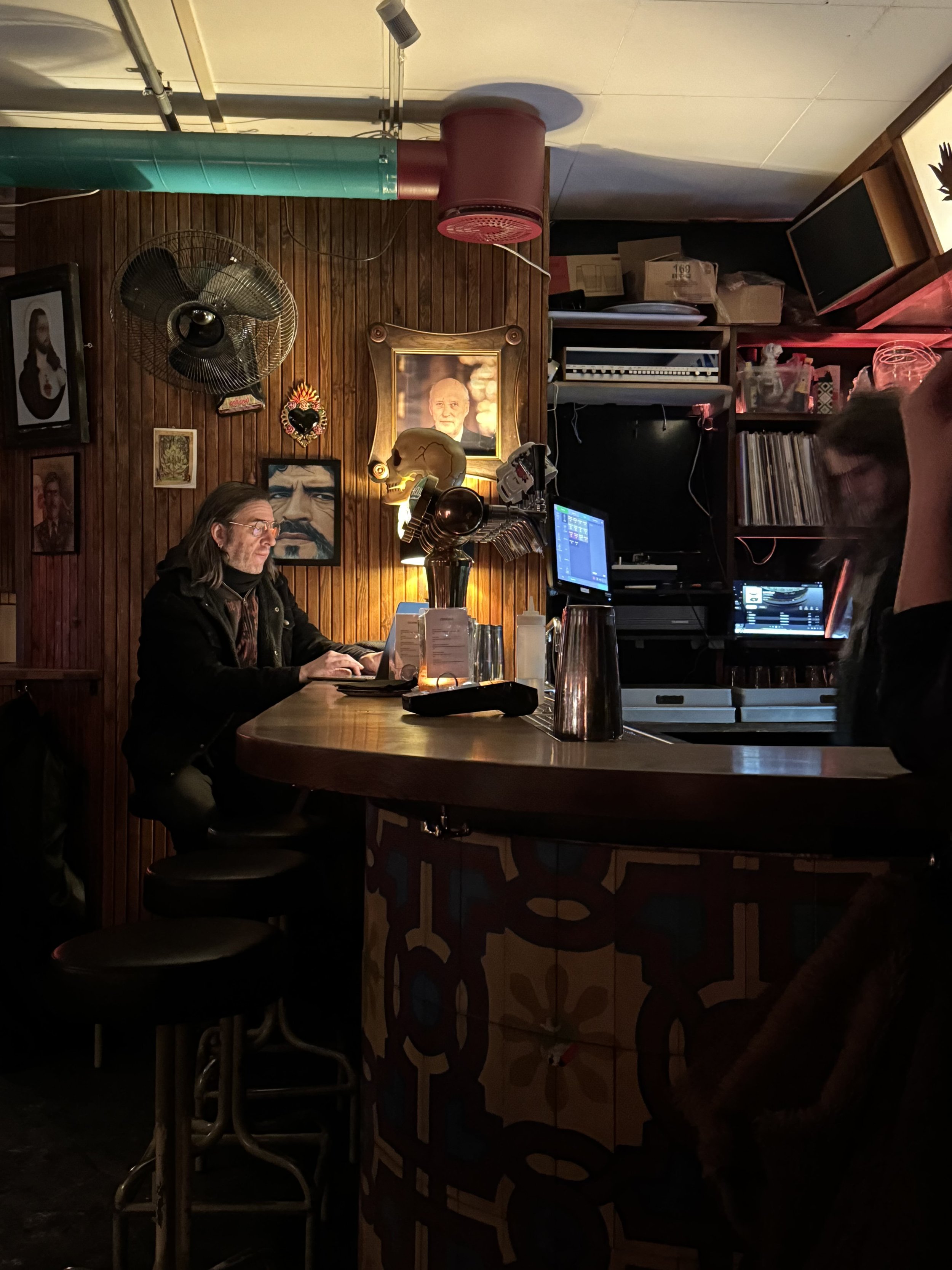 A dimly lit bar with a bartender and a customer. The bar has a wooden countertop, decorated with various pictures and a portrait of a man on the wall. There is a computer monitor and DJ equipment behind the bar. The background includes shelves with v