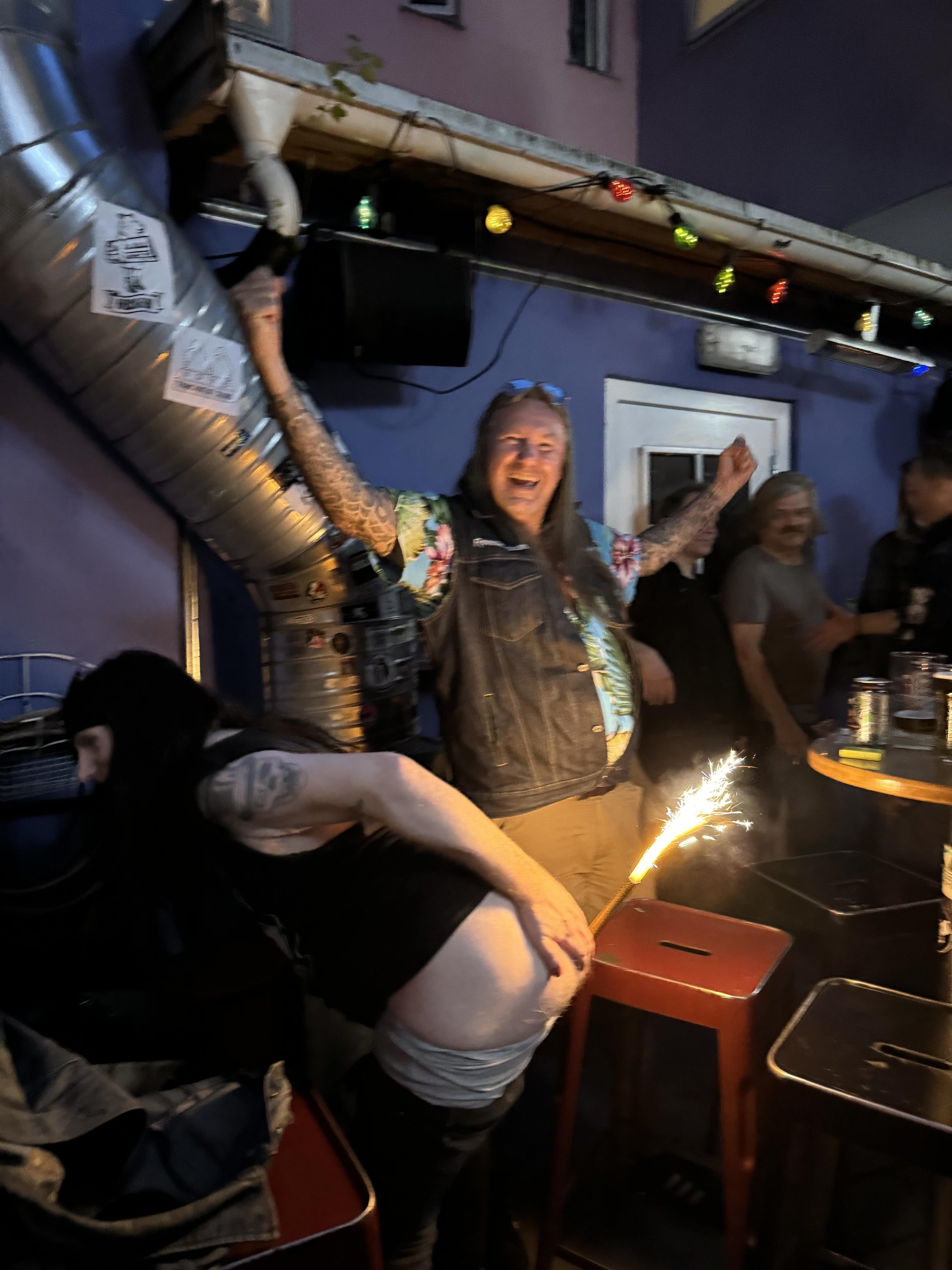 A woman with long dark hair and tattoos on her arms is bending over at a table. A woman with long, light brown hair and wearing a denim vest is standing with arms raised, smiling joyfully. There are others in the background, some with drinks, and a s